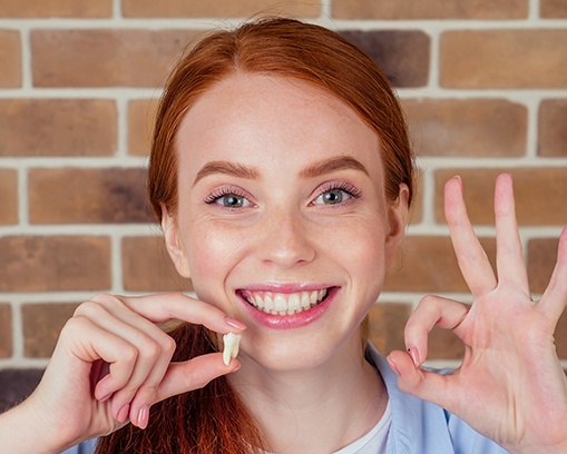 Woman with red hair holding extracted tooth in one hand and making “ok” sign with the other