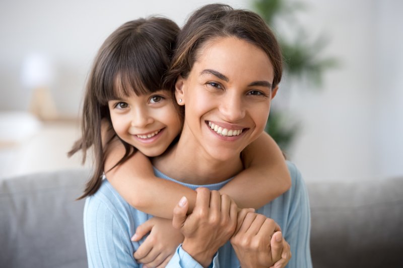 a little girl hugging a young woman from behind with her arms crossed around the woman’s neck and shoulders